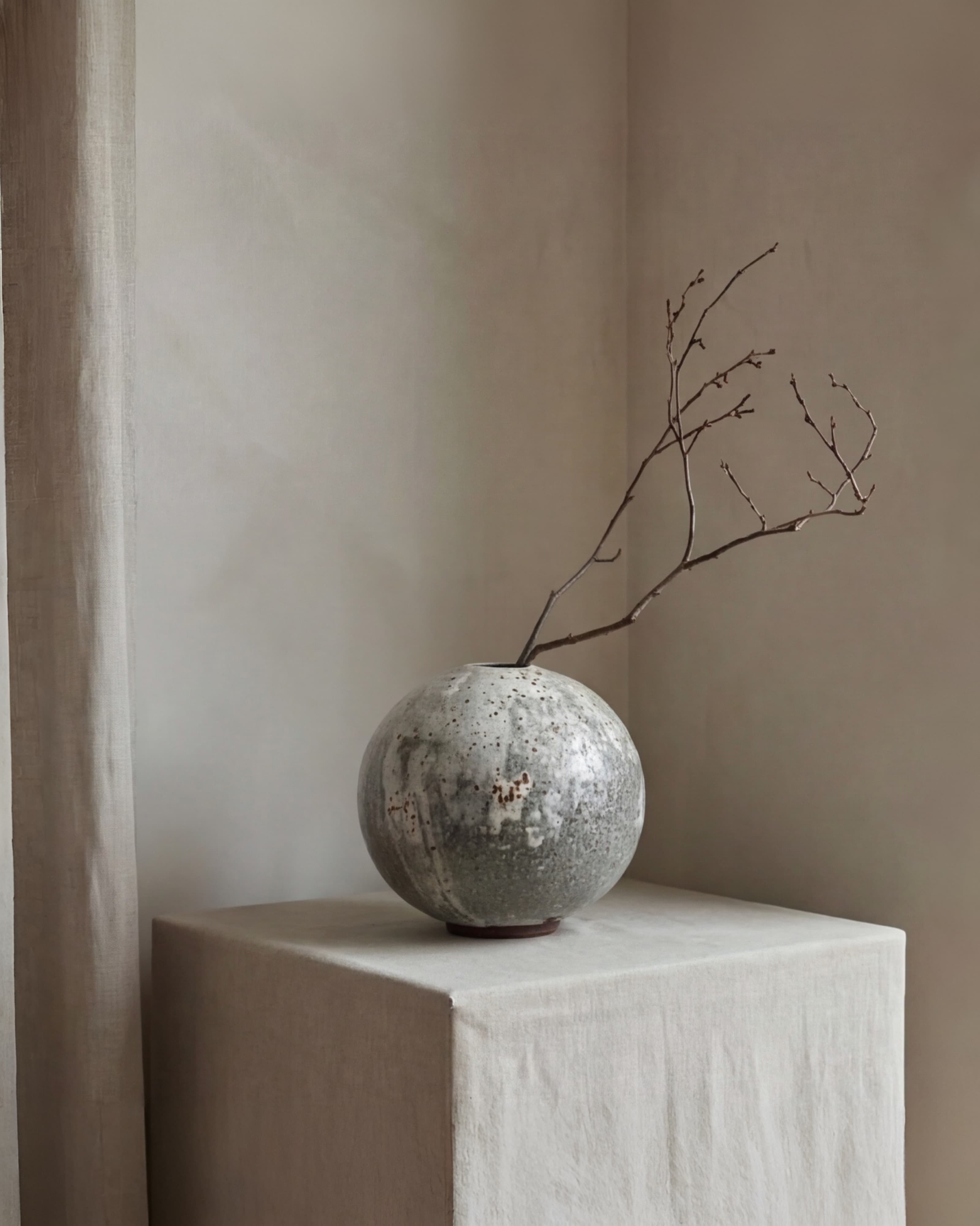 A round ceramic vase with a bare branch, resting on a linen-covered plinth